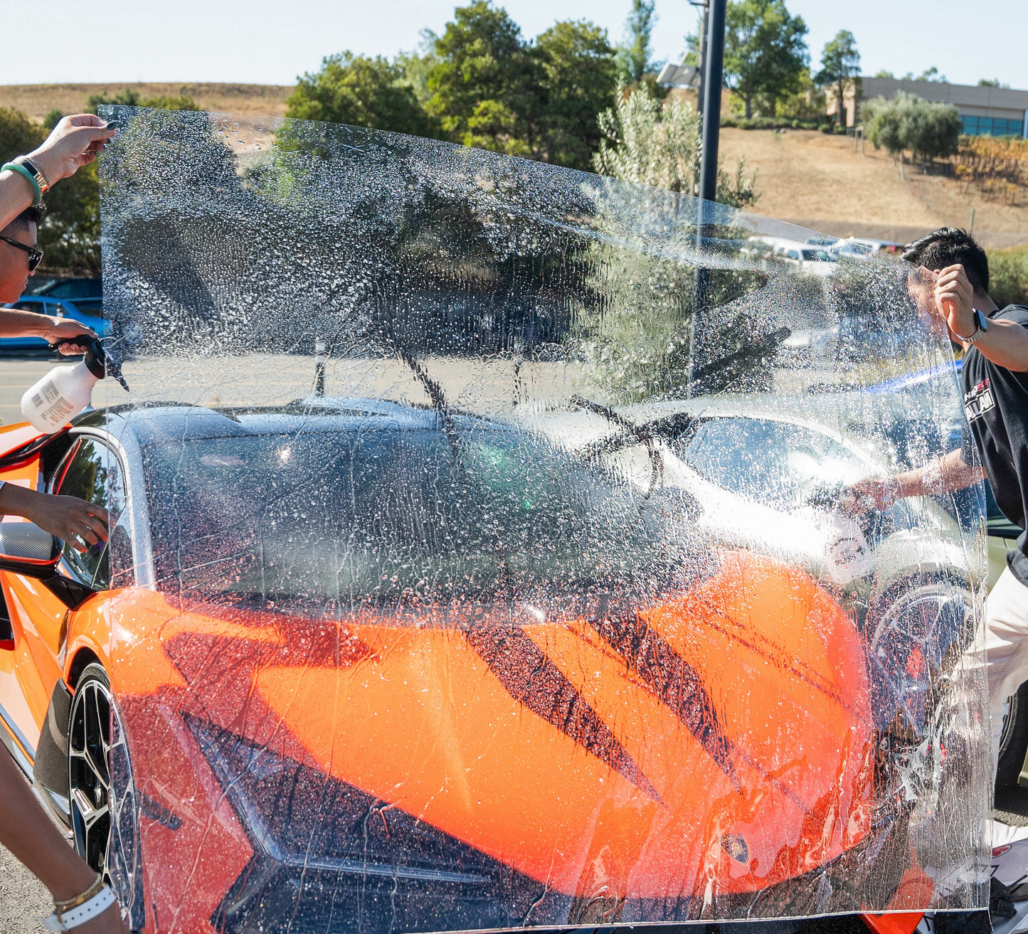 Orange sports car being washed to apply windshield film with a group of people around it.