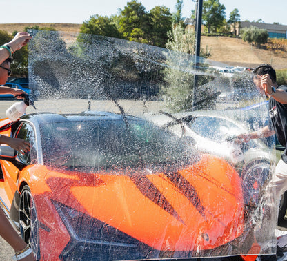 Orange sports car being washed to apply windshield film with a group of people around it.